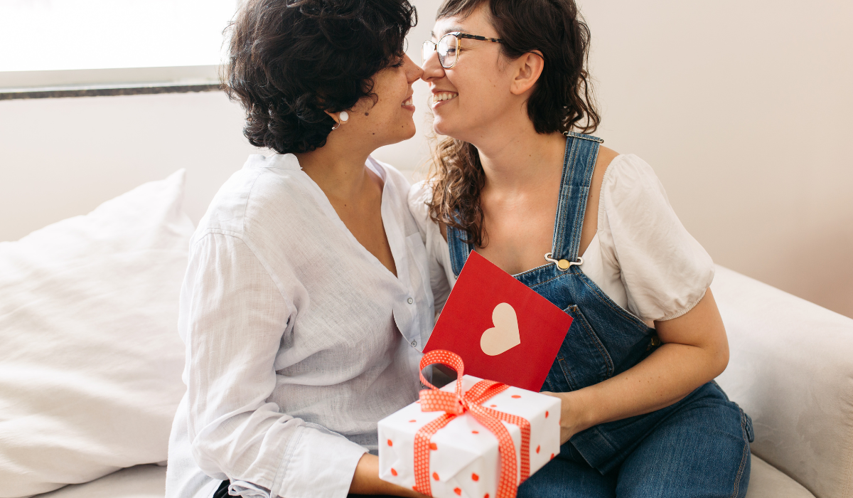 Queer couple exchanging a wrapped gift and card, celebrating love during Pride Month